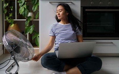 A woman working on her laptop at home while using a floor fan to stay cool.